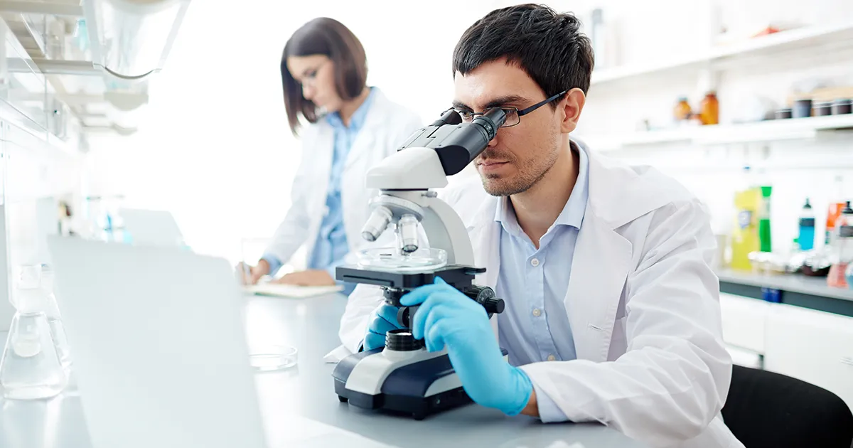 Chercheur dans un laboratoire entrain de regarder au microscope avec son assistance prenant des notes.