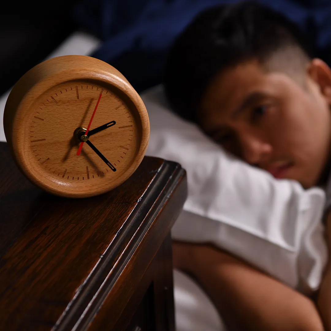 Homme asiatique dans son lit au second plan un peu flou regardant son réveil à cadrant en bois défiler à cause de son insomnie.