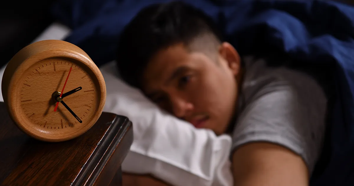 Homme asiatique dans son lit au second plan un peu flou regardant son réveil à cadrant en bois défiler à cause de son insomnie.